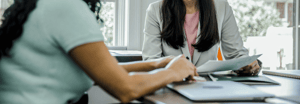 Two professionals in a meeting, reviewing documents across a desk in a bright office setting.